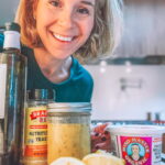 A woman smiling in a kitchen with jars of nutritional yeast, a glass jar with a sauce, miso paste, olive oil, and sliced lemons in the foreground. She has short blonde hair and is wearing a blue top.