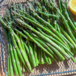 A metal tray filled with fresh green asparagus spears, placed on a wooden surface. A lemon half is partially visible on the right side. The scene is well-lit, highlighting the asparagus texture and natural color.