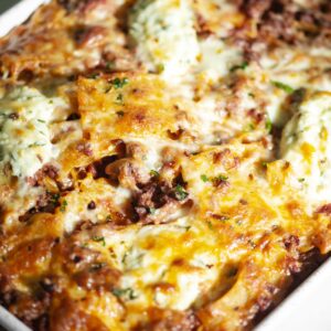 Close-up of a baked lasagna in a rectangular dish. The lasagna is golden brown on top, with visible layers of melted cheese, meat, and pasta. Garnished with parsley, it looks rich and savory.