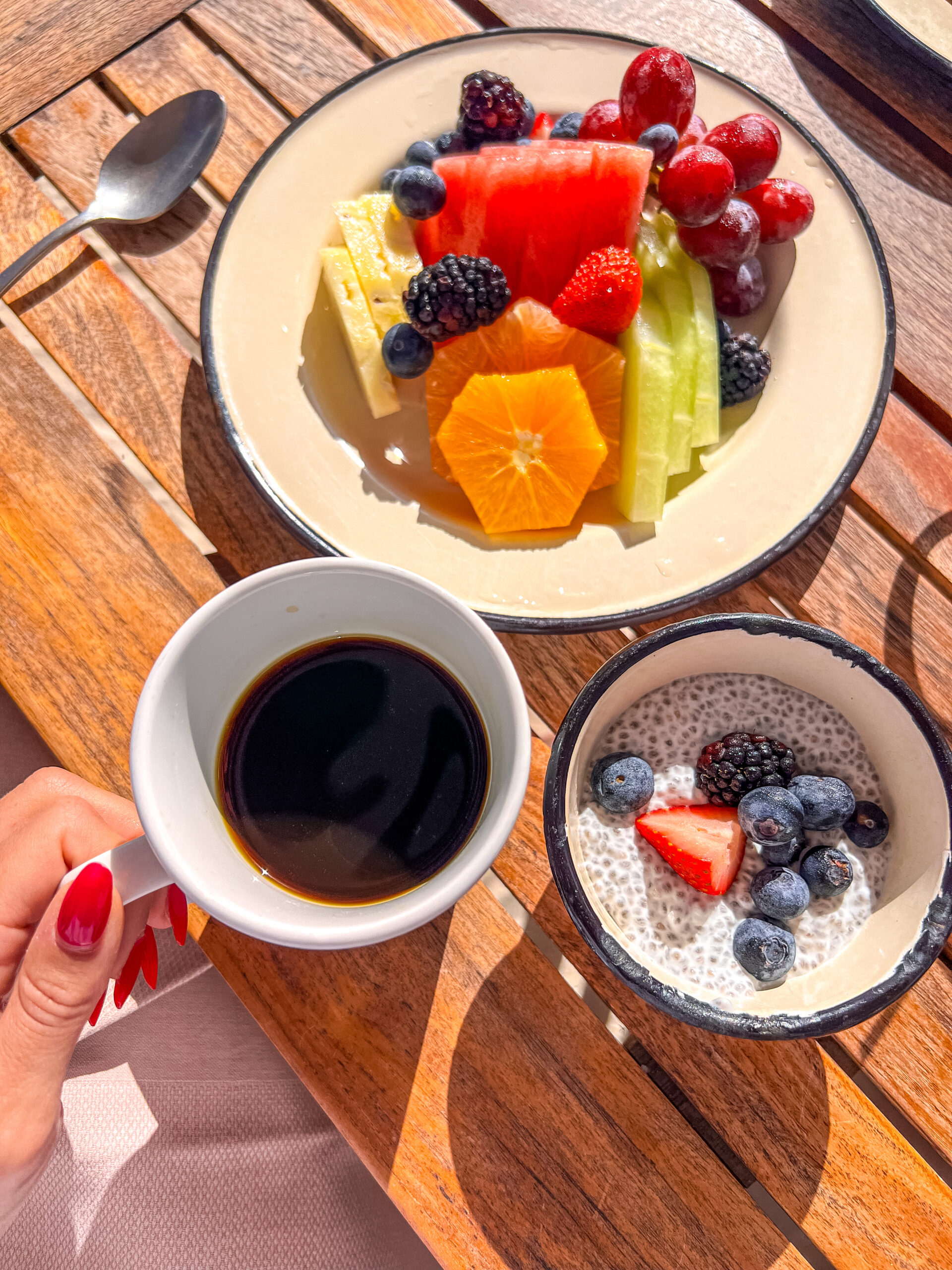 A plate of assorted fruits including watermelon, orange slices, grapes, and berries on a wooden table. Next to it is a bowl of chia pudding topped with blueberries and strawberries, and a hand holding a cup of black coffee.