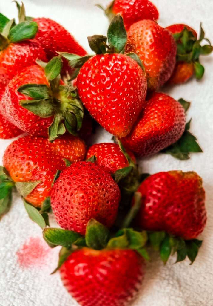 A cluster of fresh, ripe strawberries with green leaves is scattered on a white towel. The strawberries are vibrant red, indicating freshness and ripeness. One of the berries has left a small red stain on the towel.