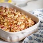 A baked casserole dish filled with a golden-brown mix of chopped ingredients, possibly bread and meat, sits on a marble countertop. Nearby are stacked plates, forks, and a patterned cloth napkin.