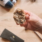 A hand holding a bitten cookie filled with chocolate chunks over a wooden floor. Nearby are white and blue shoes, mini textured golf balls, and a black pouch with gold text.