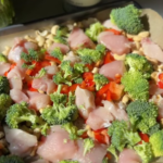 A baking tray filled with chopped raw chicken, broccoli florets, red bell peppers, and cashews, ready to be cooked. A bottle of avocado oil is visible in the background, alongside a leafy green vegetable.