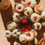 A wooden board with assorted small muffins, some topped with blueberries, strawberries, and raspberries, surrounds a bowl of syrup. Nearby are a mug, a grapefruit slice, and a plate with more muffins on a wooden table.