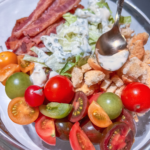 A colorful salad in a glass bowl featuring cherry tomatoes in various colors, bacon strips, lettuce, croutons, and drizzled with ranch dressing. A spoon rests in the bowl, adding a touch of silver.