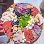 A colorful salad bowl featuring sliced pepperoni, mushrooms, green bell peppers, black olives, cherry-sized mozzarella balls, red onions, and croutons on a bed of lettuce. The bowl is set on a light surface.