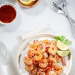 A plate of seasoned shrimp garnished with lemon wedges and parsley. Nearby are lemon slices in a bowl, a small dish of sauce, and several forks. The table surface is white, creating a clean and fresh presentation.