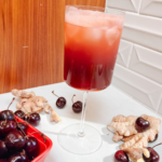 A tall glass filled with a dark red cherry and ginger juice on a white surface, surrounded by fresh cherries and ginger roots. A red basket of cherries adds to the scene. The background features white geometric tiles and wood paneling.