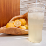 A tall glass filled with iced lemonade is on a table. Behind it, a wooden bowl holds several whole and sliced lemons. The background features wood and tiled surfaces.
