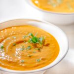Two white bowls filled with creamy orange soup are garnished with fresh green herbs. Pale textured surface in the background adds to the bright presentation.