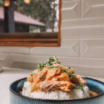 A bowl of white rice topped with shredded chicken and garnished with herbs. The dish is served in a blue bowl placed on a white counter. A window in the background reveals an outdoor view with greenery and a building.