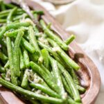 A wooden dish filled with cooked green beans topped with grated cheese. A silver fork rests beside the beans. In the blurred background, theres a stack of lemon slices on a small plate. The scene is set on a light-colored tablecloth.