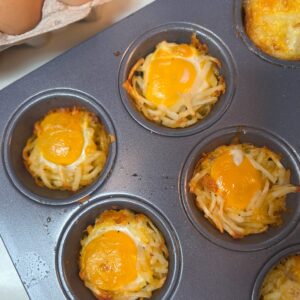 Muffin tin with hash brown and cheese breakfast cups, each topped with a sunny-side-up egg. A carton with eggs is partially visible in the background.