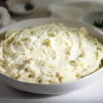 A bowl of creamy mashed potatoes garnished with herbs. The background includes a blurred glimpse of a striped napkin and a dish with additional herbs.