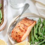 A plate with a piece of grilled salmon, seasoned and browned, accompanied by sautéed green beans sprinkled with grated cheese. Lemon wedges are placed on the side. In the background, a glass of water with a lemon slice.
