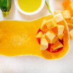 Halved butternut squash with one half cubed, surrounded by zucchini, carrots, red bell pepper, and a small bowl of olive oil on a white surface.