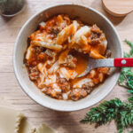 A bowl of lasagna soup with pasta, ground meat, and herbs in a light-brown broth. A red-handled spoon rests inside the bowl. Nearby are uncooked lasagna sheets and sprigs of fresh herbs on a wooden surface.