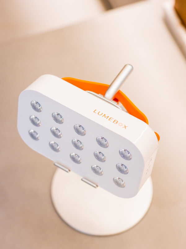 Close-up of a white LumiBox device with multiple small, round lights on its surface. The device is mounted on a silver stand, with an orange handle visible behind it, set against a neutral background.