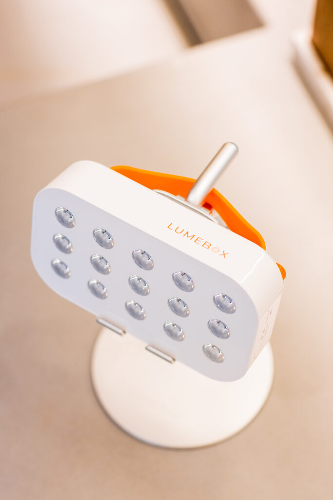 Close-up of a white LumiBox device with multiple small, round lights on its surface. The device is mounted on a silver stand, with an orange handle visible behind it, set against a neutral background.