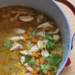 A pot of chicken soup with chunks of chicken, sliced carrots, and celery in broth. Garnished with fresh parsley. The pot has a red handle and rests on a wooden surface.