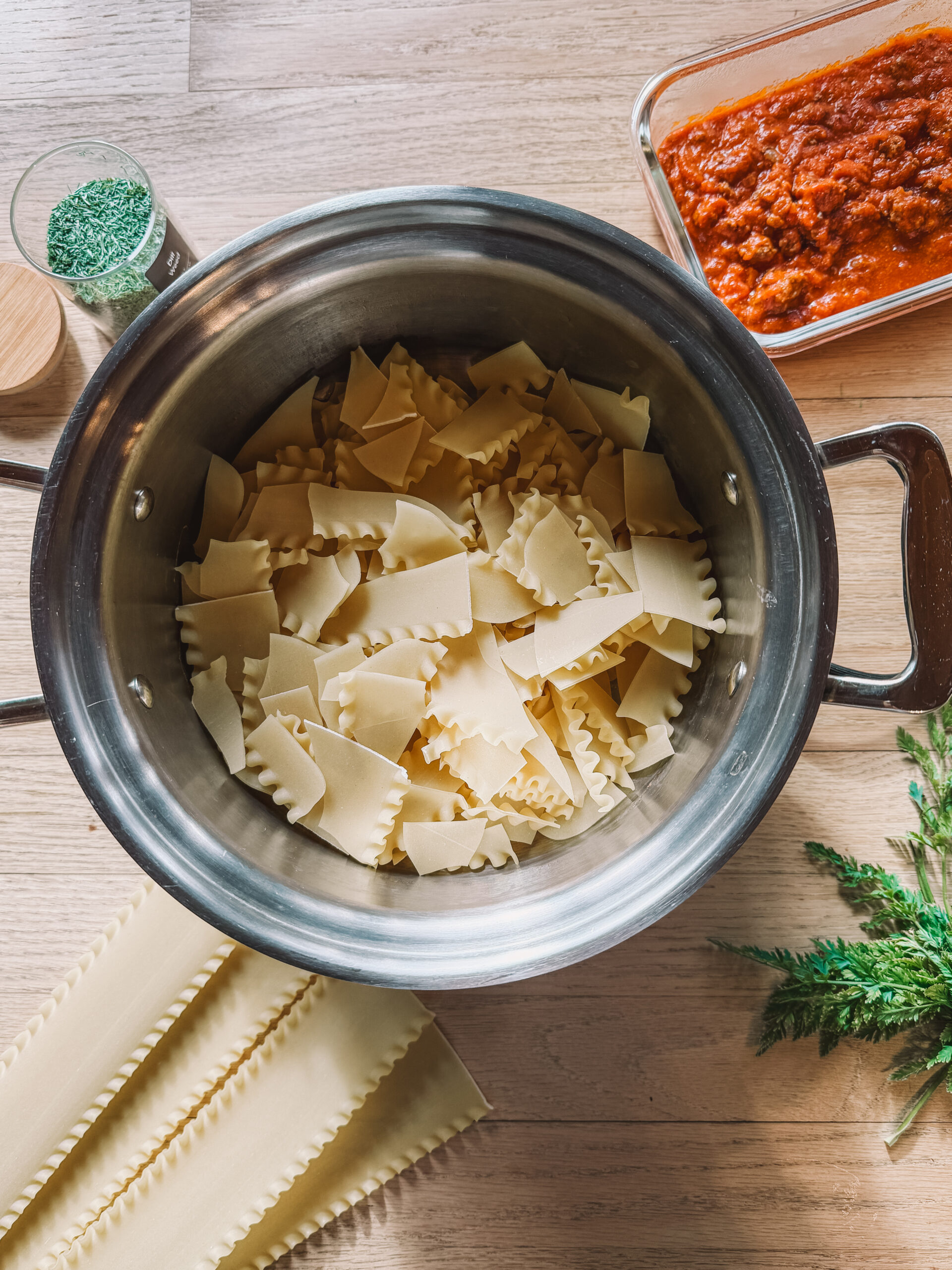 A top view of a kitchen setup featuring a stainless steel pot filled with pieces of uncooked lasagna sheets. Nearby, theres a rectangular dish with red tomato sauce, a jar of herbs, and some fresh green herbs on a wooden table.