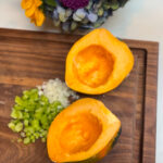 A halved acorn squash is on a wooden cutting board with chopped celery and onions. In the background, there is a vibrant bouquet of flowers with red roses, purple chrysanthemums, and yellow sunflowers.