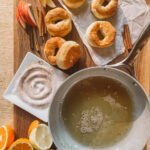 Wooden board with several air fryer apple cider doughnuts on parchment paper, sliced apples, cinnamon sticks, a citrus assortment, and a deep pan filled with oil. A plate with cinnamon sugar is nearby, ready for doughnut coating.