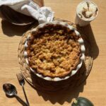 A freshly baked pie with a crumbly topping sits on a woven mat. Nearby are a spoon, fork, an acorn squash, and a cup of ice cream with chocolate swirls on a wooden table. A wooden lid and a white cloth are in the background, all bathed in sunlight.