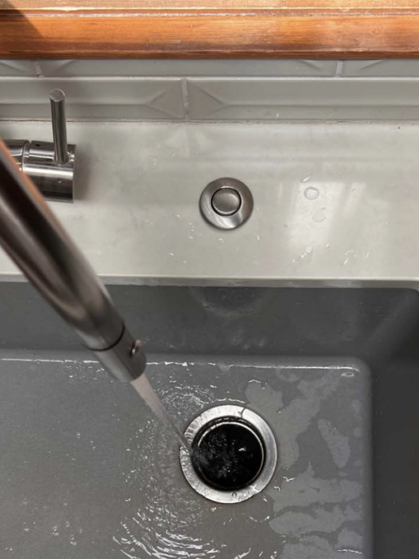A stainless steel kitchen sink with water running from a modern faucet into the drain. The countertop is gray, with a wooden border at the top. A soap dispenser is installed beside the faucet, and the wall has white tile backsplash.