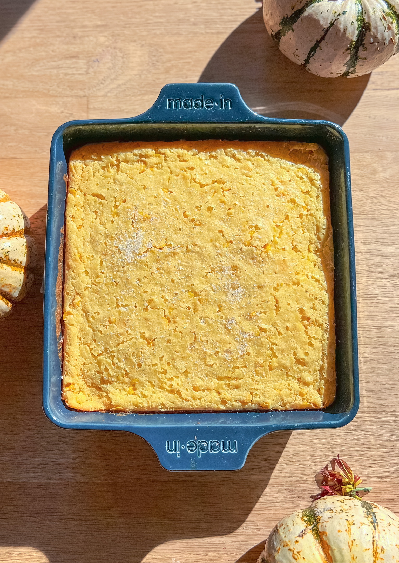 A blue square baking dish holds a golden brown cornbread, placed on a wooden surface. The dish has made-in embossed on its handles. Decorative gourds surround the dish, adding an autumnal touch.