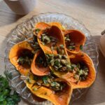 A glass plate with roasted butternut squash slices topped with herbs and seeds on a wooden table. A lit candle is in the background, and a sprig of parsley is on the left side.