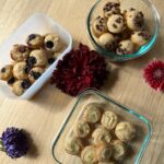 Three containers of assorted mini muffins are arranged on a wooden table, surrounded by colorful flowers. One container holds blueberry muffins, another has chocolate chip muffins, and the third contains plain muffins.