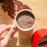 A hand holding a mug filled with frothy hot chocolate. Cocoa powder is scattered on the wooden table. A red pot with a wooden handle next to the mug is partially visible, also containing hot chocolate. Sunlight casts soft shadows.