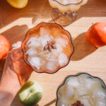 Three glasses filled with iced apple cocktails garnished with star anise are arranged on a wooden table. Several apples surround the glasses, adding color and texture to the composition, while sunlight casts soft shadows. A hand holds one glass.