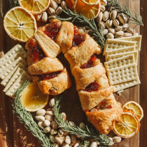 A pastry shaped like a candy cane, filled with red jam, is surrounded by sliced oranges, pistachios, crackers, and sprigs of rosemary on a wooden board. Whole oranges and crackers are also arranged around the centerpiece.