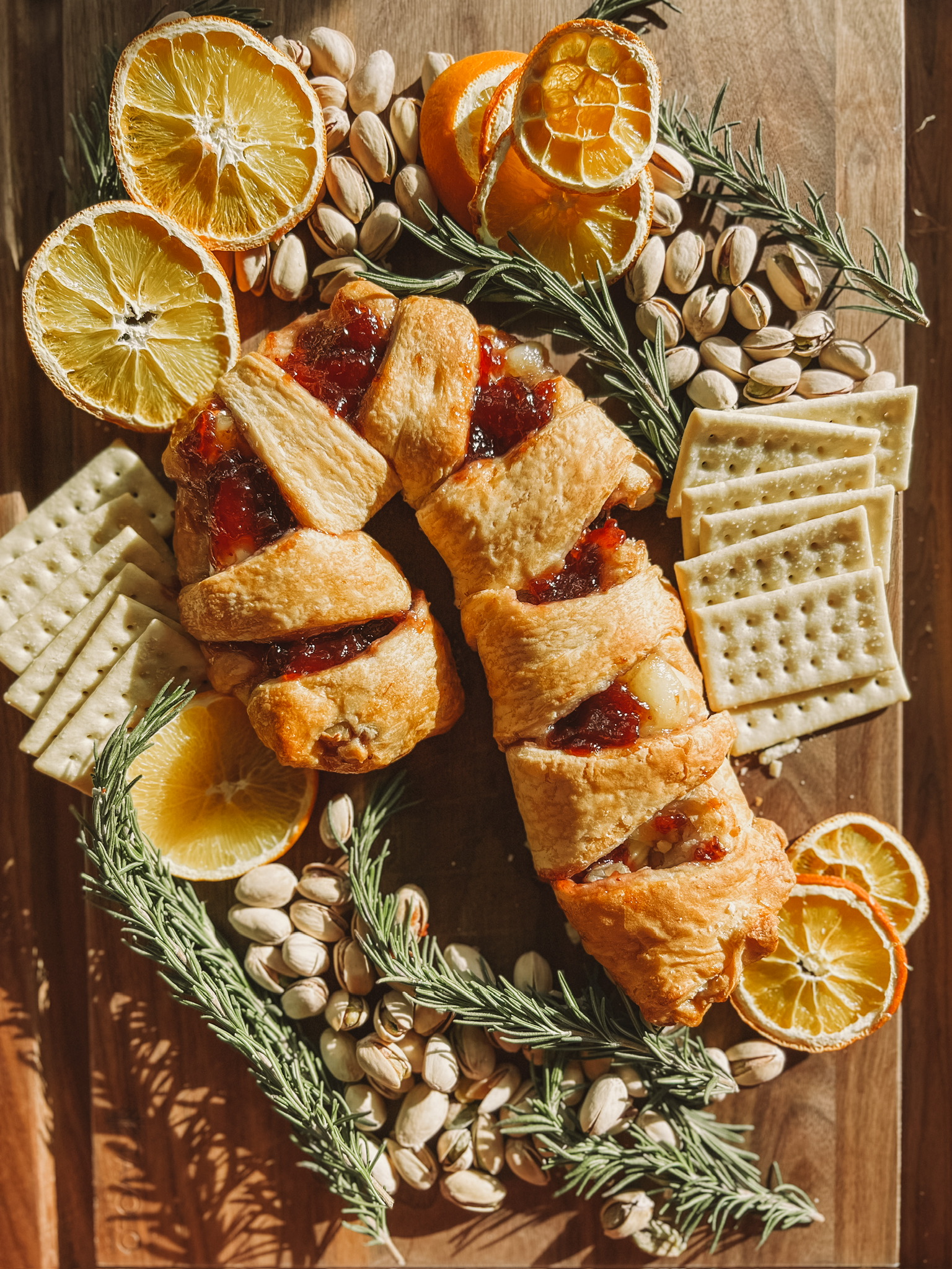 A pastry shaped like a candy cane, filled with red jam, is surrounded by sliced oranges, pistachios, crackers, and sprigs of rosemary on a wooden board. Whole oranges and crackers are also arranged around the centerpiece.
