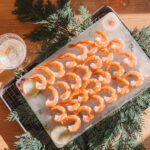 A tray of shrimp cocktails arranged on ice, garnished with lemon slices and red berries, surrounded by evergreen branches. Two cocktail glasses are placed nearby on a wooden surface.