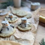Decorative cookies with melted snowman designs featuring marshmallow heads and icing faces, lie on parchment paper. A lit candle and some greenery are in the background, creating a cozy atmosphere.