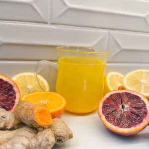 A glass mug filled with bright orange juice is surrounded by sliced citrus fruits, including oranges and lemons. Fresh ginger root and a piece of turmeric are placed alongside against a white tiled background.