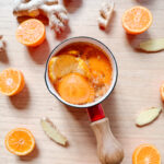 A small saucepan with orange slices and liquid sits on a wooden table, surrounded by halved and whole oranges and ginger slices. The saucepan has a red handle and the scene suggests a fresh, citrusy preparation.