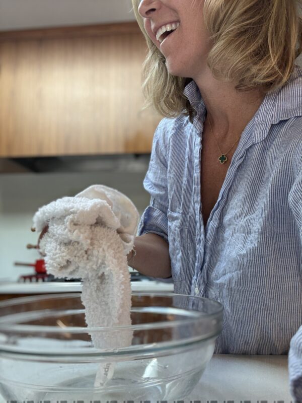 A woman smiling while wringing a white towel over a clear glass bowl in a kitchen. She is wearing a blue and white striped shirt and a clover necklace.