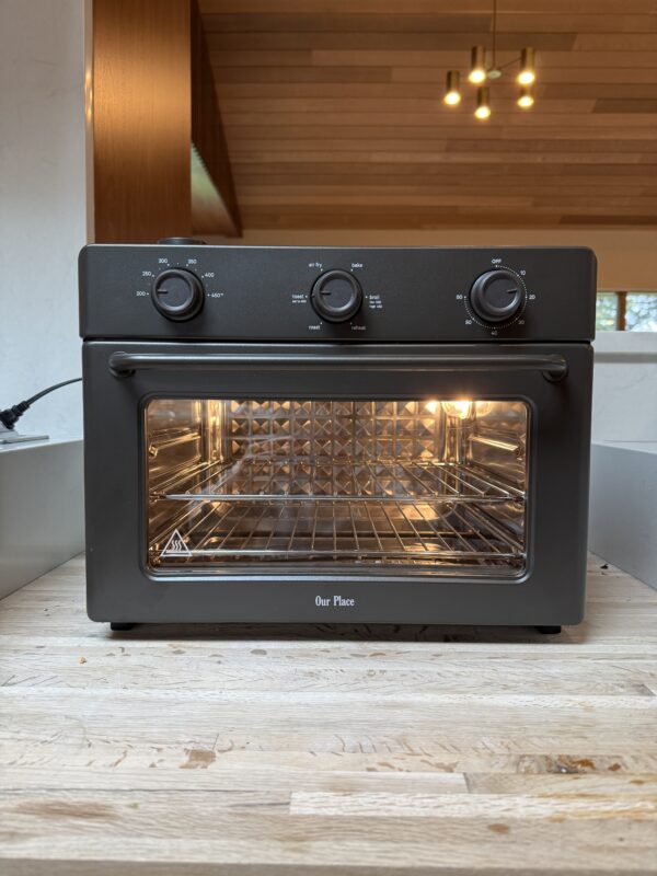 A black countertop oven with three control knobs sits on a wooden surface. The oven door is closed with the interior light on, showing wire racks inside. The brand Our Place is visible on the front.