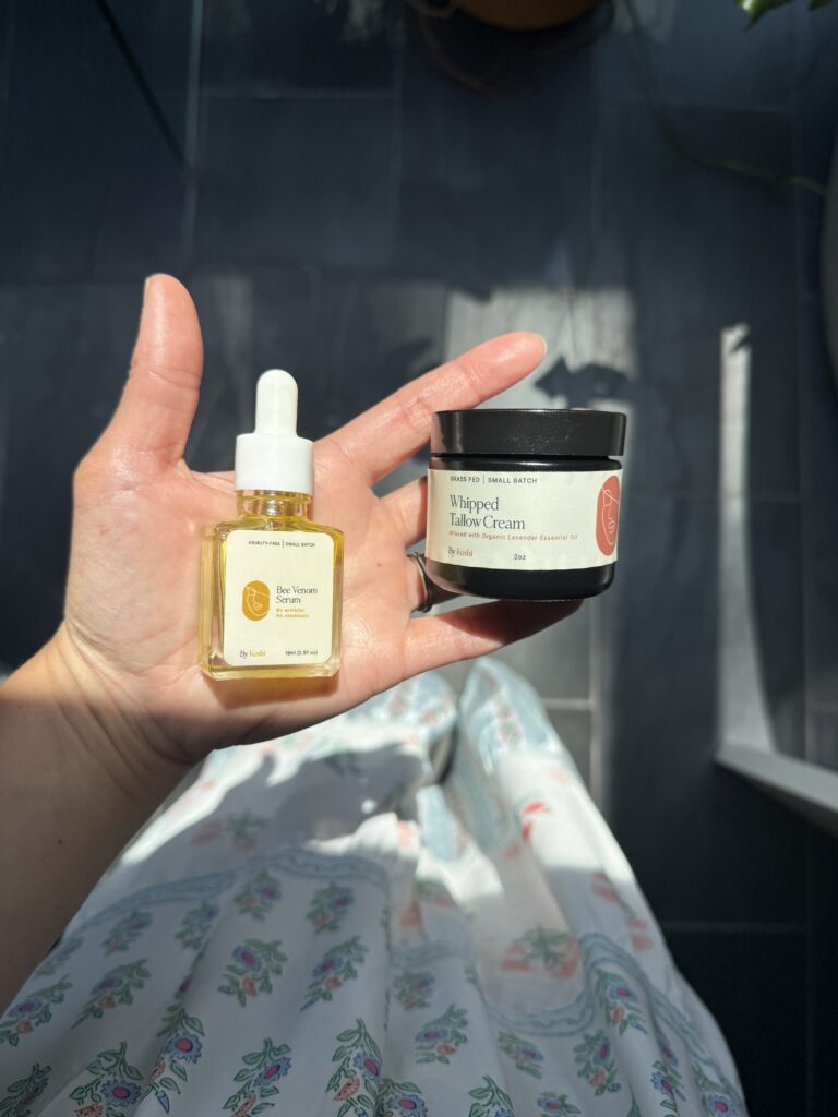 A hand holding a dropper bottle labeled Bee Venom Face Serum and a jar labeled Whipped Tallow Cream, with sunlight casting shadows and a floral skirt visible in the background.