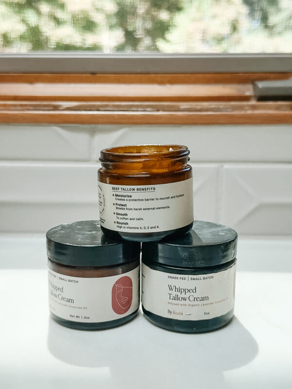 Three jars of whipped tallow cream are stacked on a white surface in front of a window. The top jar is open, revealing cream inside, while the two jars below are closed and labeled “Whipped Tallow Cream.”.