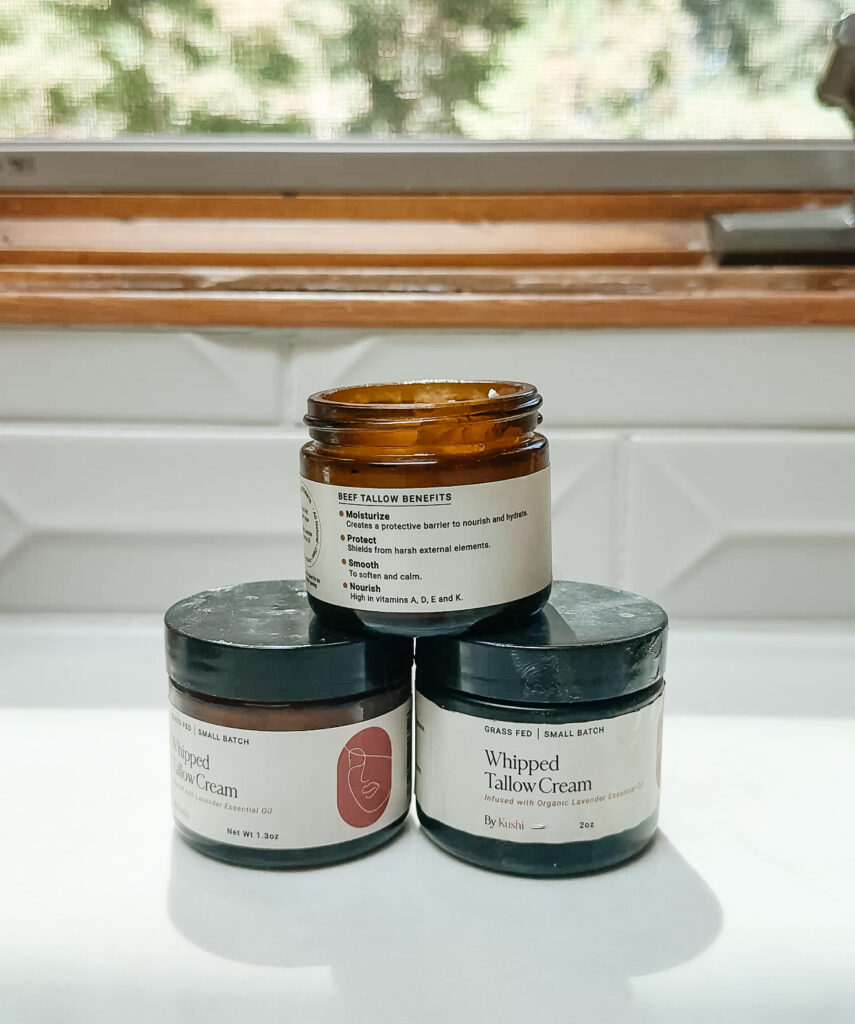 Three jars of whipped tallow cream are stacked on a white surface in front of a window. The top jar is open, revealing cream inside, while the two jars below are closed and labeled &ldquo;Whipped Tallow Cream.&rdquo;.