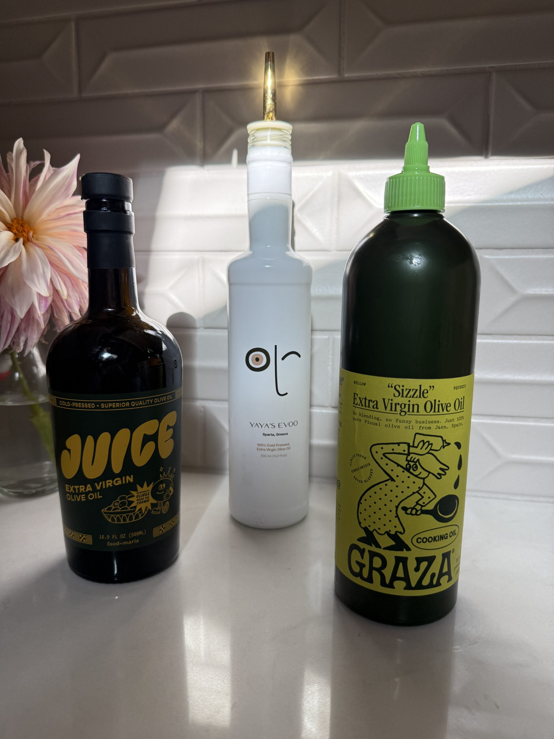 Three bottles of extra virgin olive oil are on a white kitchen counter with a geometric tile backsplash. The brands shown are Juice, Yaya&rsquo;s EVOO, and Graza &ldquo;Sizzle.&rdquo; A vase with pink flowers is in the background.