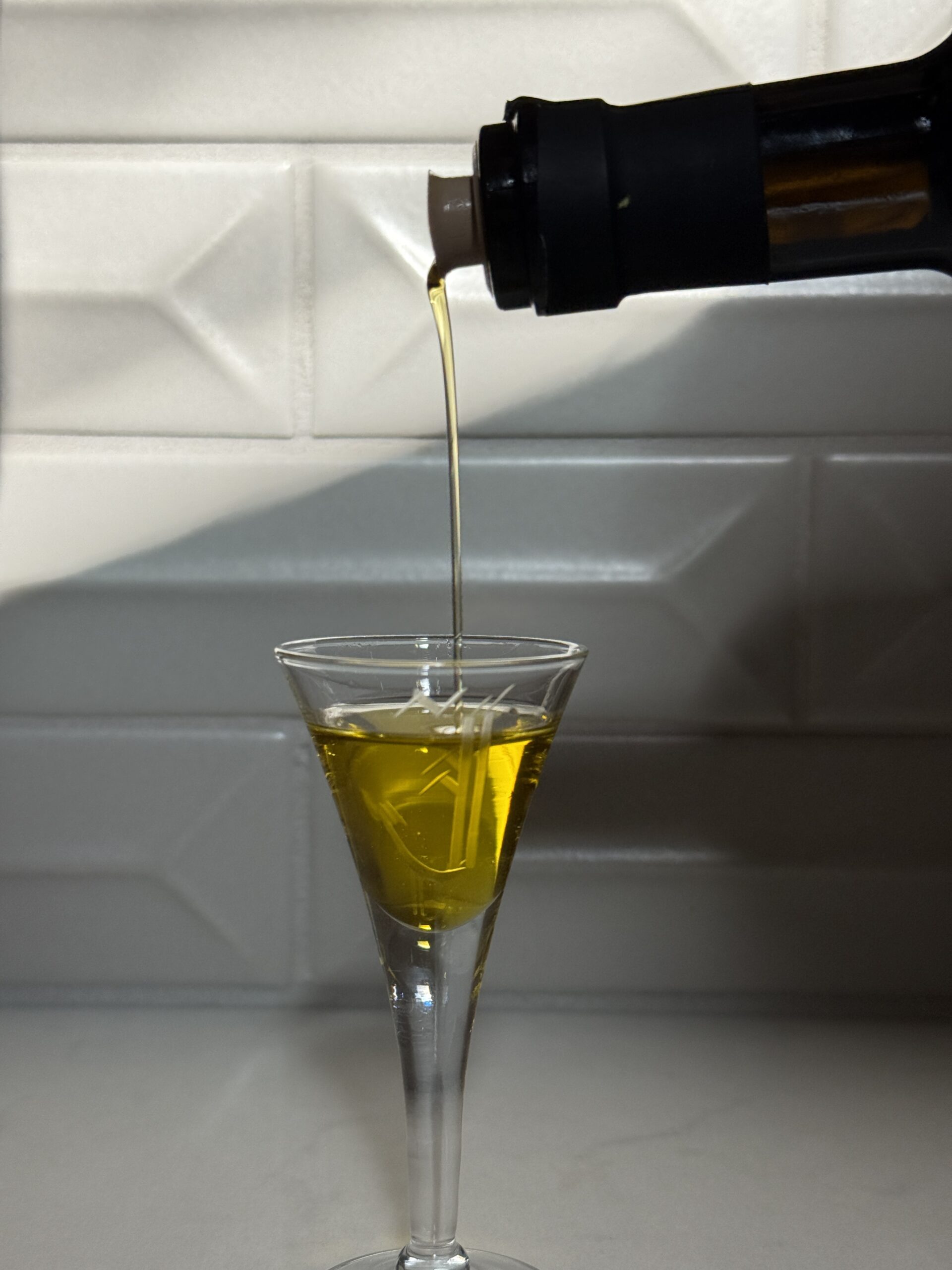 A stream of golden olive oil is being poured from a bottle into a clear, stemmed glass, set against a white tiled background with sunlight casting shadows.