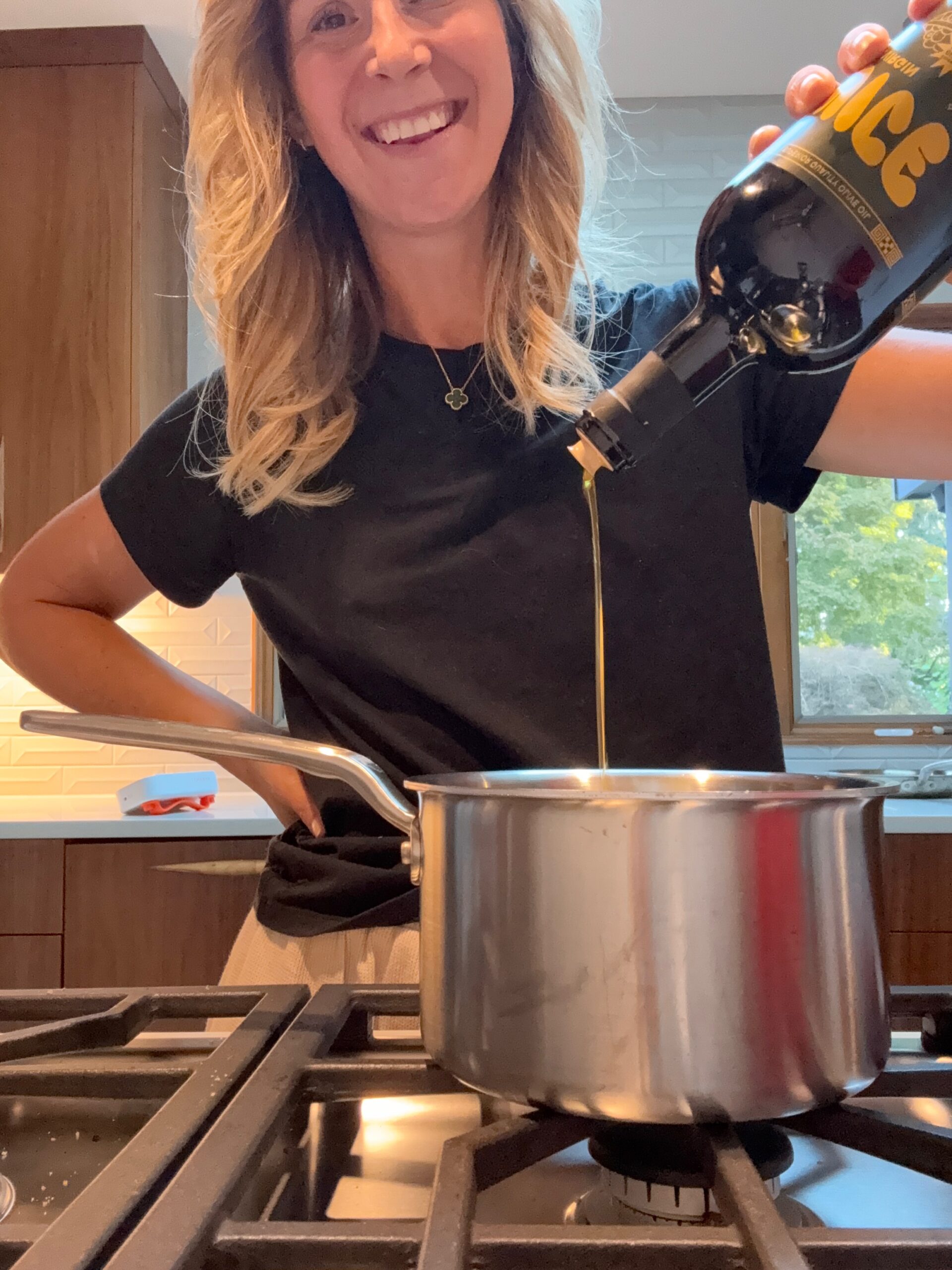 A woman smiling while pouring oil from a bottle into a pot on a stove in a modern kitchen.