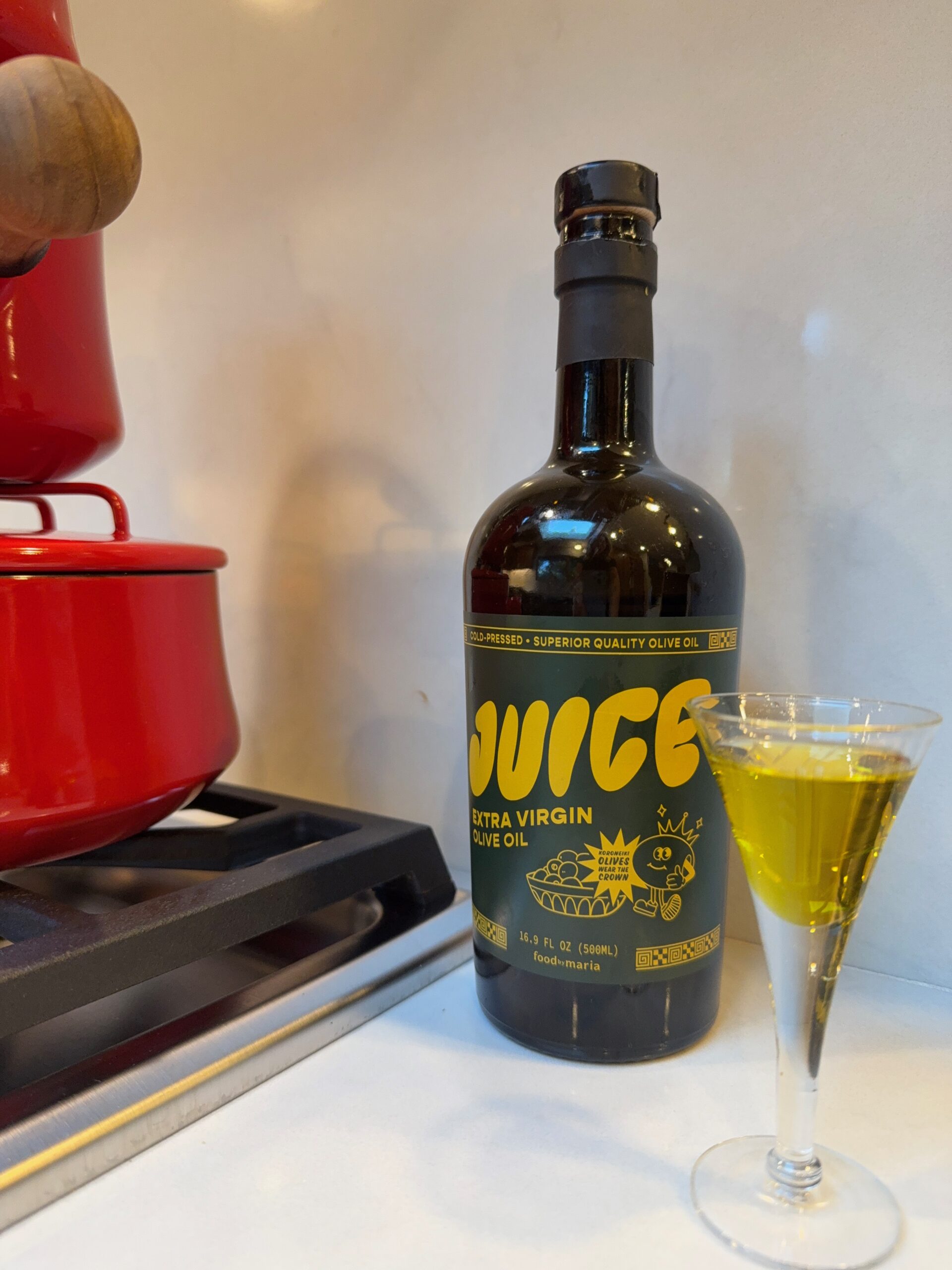 A bottle of JUICE extra virgin olive oil stands on a kitchen counter beside a red pot on a stove, with a small glass filled with olive oil in front of the bottle.